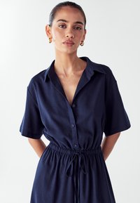 Jūrų laivyno marškinių suknelė su trumpomis rankovėmis, apykaklės dizainu, priekyje mygtukai, elastinga juosmens dalis ir surišimo detalė. Lygus paviršius, laisvas siluetas.