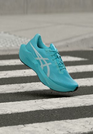 Chaussure de course bleue avec logo blanc en l'air au-dessus d'un passage piéton rayé sur une route en asphalte.