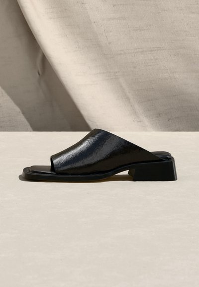 Μαύρο mule σανδάλι με ανοιχτά δάχτυλα, χαμηλό τετράγωνο τακούνι και γυαλιστερό φινίρισμα, εμφανίζεται σε προφίλ από την πλευρά πάνω σε μπεζ επιφάνεια με υφασμάτινο φόντο.