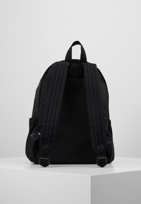 Schwarzer Rucksack mit gepolsterten, verstellbaren Schulterriemen und einem Tragegriff, präsentiert auf einem weißen Podest vor einem schlichten grauen Hintergrund.