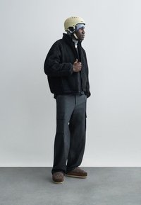 Zwarte fleece jas, grijze cargobroek, beige helm, bruine schoenen met patroon. De outfit heeft een ontspannen pasvorm en gelaagde texturen.