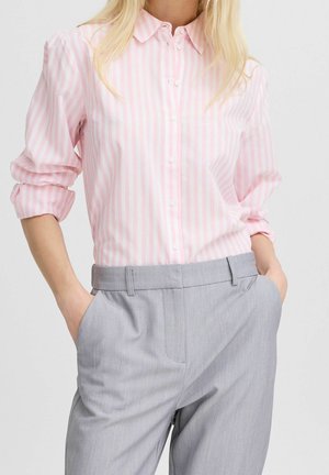 Kobieta ubrana w różowo-białą koszulę w paski z podwiniętymi rękawami, włożoną w jasnoszare, dopasowane spodnie, z rękami w kieszeniach.