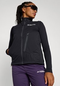 Black Terrex vest features a high collar, zippered pocket, and geometric patterns on the lower front. Paired with purple pants, highlighting athletic design.