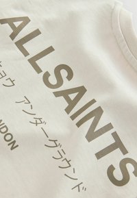 Beige mikina s potiskem "ALLSAINTS" v tmavě olivové zelené. Má kulatý výstřih a měkkou texturu tkaniny. Obsahuje také další text v japonštině.