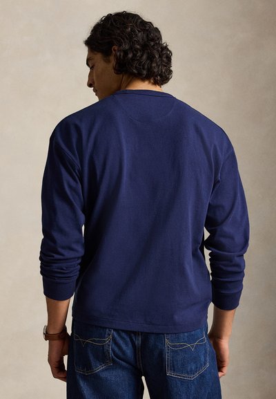 Άνδρας με σγουρά μαλλιά με την πλάτη γυρισμένη, φορώντας μια μπλε μπλούζα με μακριά μανίκια και μπλε τζιν, στέκεται μπροστά σε ουδέτερο φόντο.