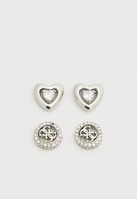 Boucles d'oreilles ton argent avec un dessus en forme de cœur serti d'une pierre claire et une base circulaire ornée de cristaux transparents et d'un logo.