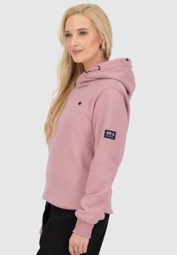 Donna bionda che indossa un felpa rosa con una toppa sulla manica, abbinata a pantaloni neri, di profilo a sinistra su uno sfondo semplice.