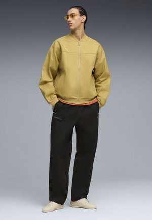 Uomo con giacca gialla senape con cerniera, pantaloni neri con loghi, sneakers beige e occhiali con lenti gialle, in piedi con le mani nelle tasche.