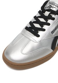 Strieborná športová teniska s lesklým syntetickým vrškom, čiernymi šnúrkami, vystlaným golierom a gumovou podrážkou s hnedými akcentmi. Na boku je logo.