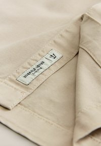 Tissu beige avec une texture lisse, comportant une étiquette indiquant "DURABLE GOODS" et un logo de couronne. Pliée, mettant en valeur les détails de couture.