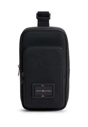 Kompakt sort crossbody-taske med lynlåslomme foran, stofstruktur og Tommy Hilfiger-logoet placeret centralt under lommen.