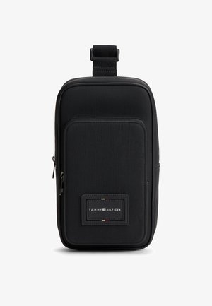 Kompakt sort crossbody-taske med lynlåslomme foran, stofstruktur og Tommy Hilfiger-logoet placeret centralt under lommen.