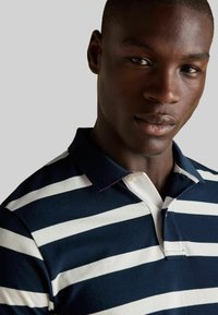 Jūras spēku un baltas svītrainas polo krekls ar apkakles dekoltē un divu pogu aizdari, ar faktūrētu audumu un kontrastējošu apkakles detaļu.