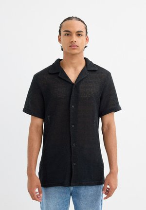 Camisa negra de manga corta y cierre de botones, hecha de tejido texturizado y ligero, con cuello. Presenta un ajuste holgado y detalles de costura visibles.