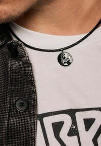 Collana in corda nera intrecciata con un pendente circolare in argento che presenta un design di uno scorpione, indossata sopra una maglietta chiara e una giacca stampata.