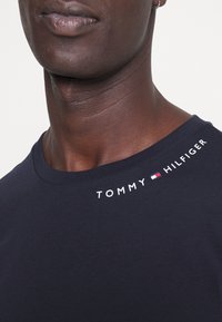 Tmavě modré bavlněné tričko s kulatým výstřihem, na levém rameni bílé logo „TOMMY HILFIGER“. Hladká textura, přiléhavý střih.