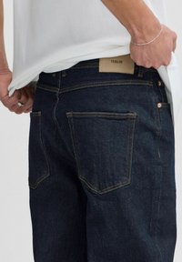 Dark blue denim jeans with a tan leather patch at the waist, copper rivets, and standard back pockets. The fabric has slight texture.