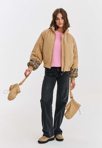 Žena v béžovej prešívanej bunde s leopardími manžetami, ružovom svetri, čiernych džínsoch a béžových topánkach, drží zodpovedajúce béžové rukavice na popruhoch.