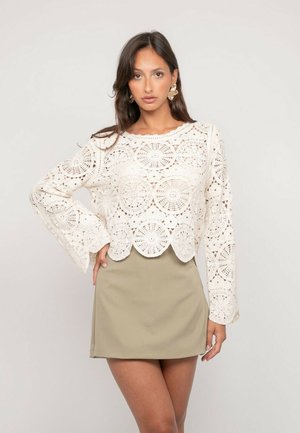 Femme portant un haut blanc en crochet à manches longues et une mini-jupe beige, posant avec une main sur la hanche devant un fond uni.