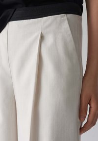 Pantaloni plissettati di colore bianco sporco realizzati in un tessuto testurizzato, con una cintura nera a contrasto. Mano appoggiata di lato.