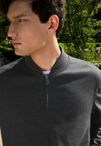 Dunkelgraue Bomberjacke mit glatter Textur, geripptem Kragen und Reißverschlussdetails vorne, getragen über einem weißen Shirt. Außenaufnahme mit Grünpflanzen im Hintergrund.