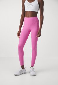 Roze visokotrajne tajice s glatkom teksturom, s bočnim džepovima i Nike logotipom. Uparene s bijelim športskim cipelama i odgovarajućim gornjim dijelom.