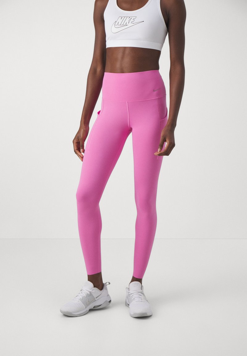 Rosa hochtaillierte Leggings mit einer glatten Textur, ausgestattet mit Seitentaschen und einem Nike-Logo. Kombiniert mit weißen Sportschuhen und einem passenden Oberteil.