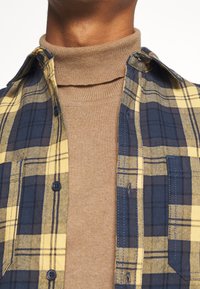 Camisa de franela a cuadros azul y amarilla, superpuesta sobre un cuello alto beige. Presenta cierres de botón y texturas contrastantes en la tela.