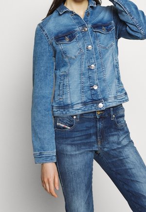Femme portant une veste en jean de lavage moyen avec des boutons argentés et un jean bleu assorti sur un fond blanc uni.
