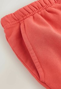 Krátke nohavice z jemnej koralovej látky s elastickým pásom a stranovým vreckom, zobrazené na svetlom pozadí.