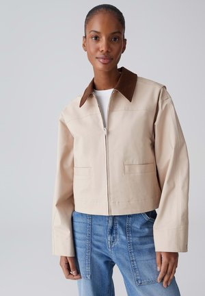 Frau trägt eine beige Jacke mit Reißverschluss, einem dunkelbraunen Kragen und hellblauen Jeans mit aufgesetzter Tasche, steht vor einem schlichten hellgrauen Hintergrund.