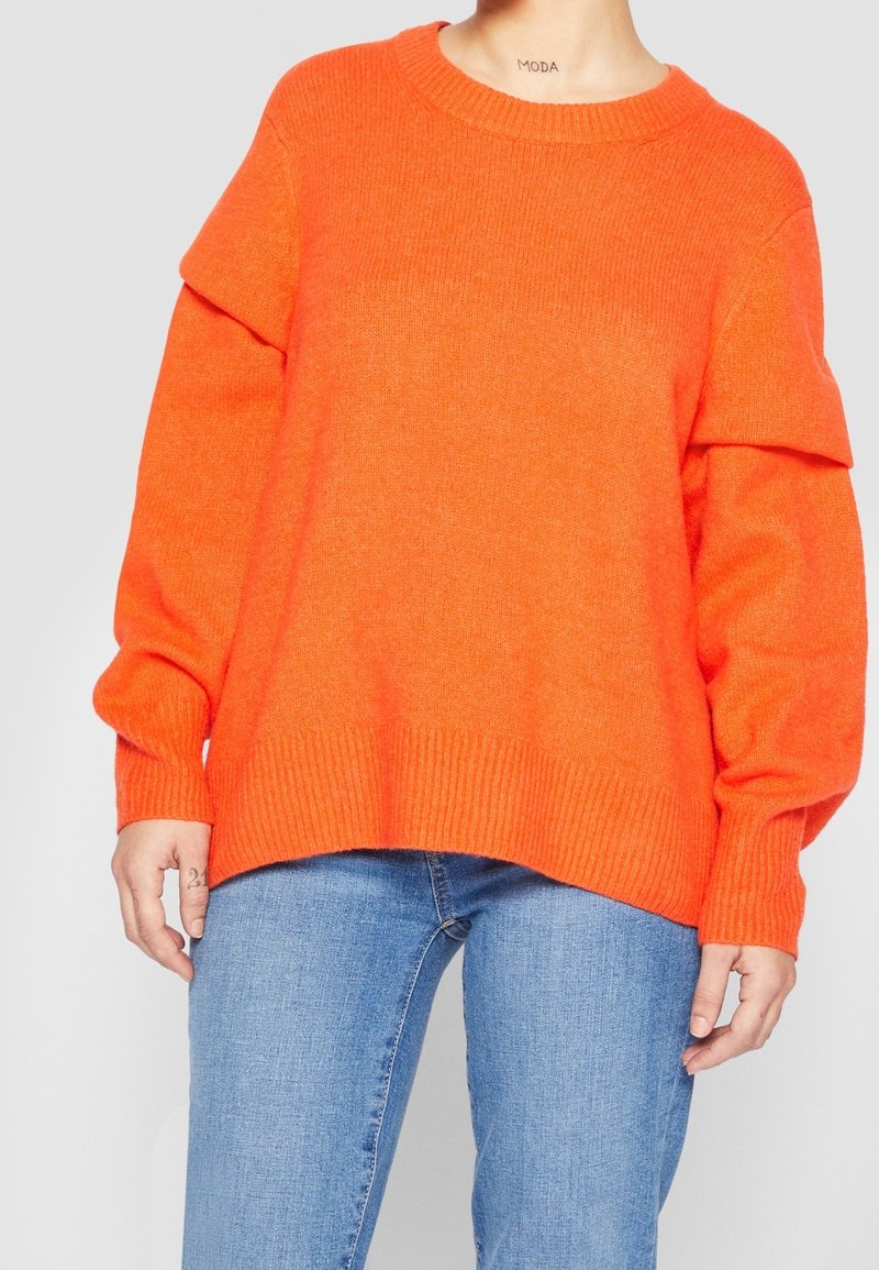 Personne portant un pull orange vif à manches longues et un jean bleu, debout devant un fond gris clair uni.