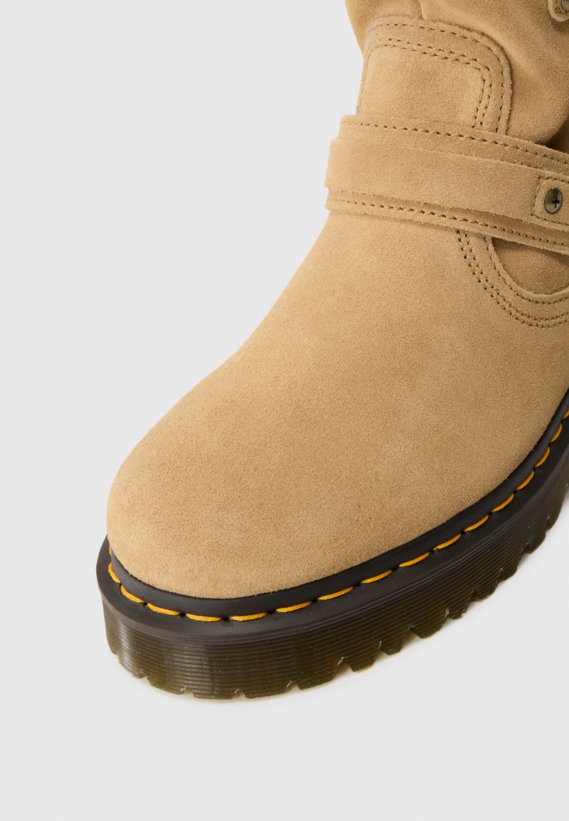 Stivaletto alla caviglia in suede beige con punta rotonda, dotato di una suola nera spessa e dettaglio di cucitura gialla. Include una fascia con un bottone.