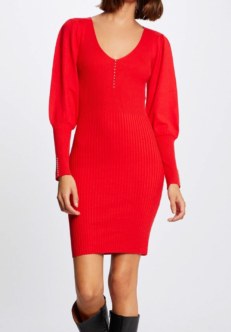 Femme portant une robe en maille côtelée rouge près du corps avec de longues manches bouffantes, un col en V, des boutons décoratifs et des bottes noires montantes jusqu'au genou.