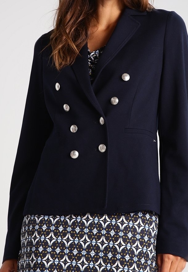 Blazer doppiopetto blu navy con bottoni in metallo, rever a lancia e due tasche frontali, abbinato a una gonna fantasia con disegni geometrici.