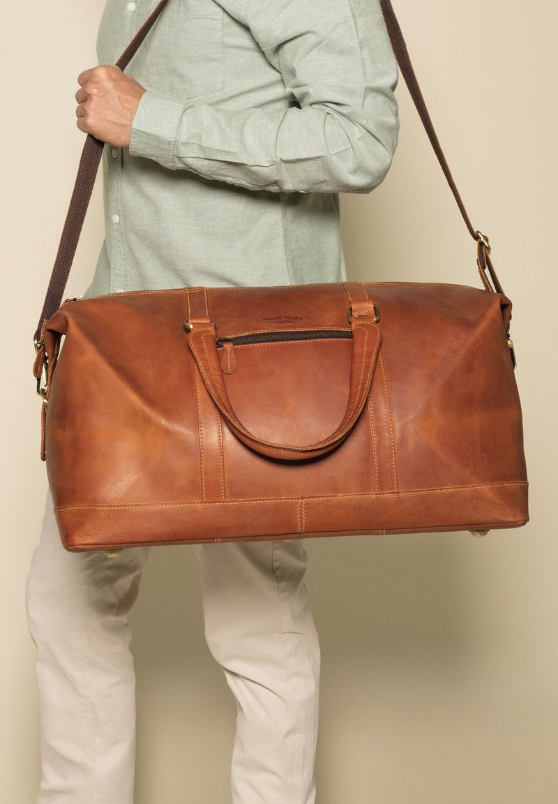 Bruine leren reisbag met dubbele handgrepen, een ritsvak en een bruine schouderriem. Gladde textuur en rechte vorm.