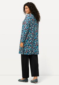 Túnica floral en azul, con una variedad de colores que incluyen flores rosas y blancas. Combinada con pantalones negros sueltos y zapatos azules.