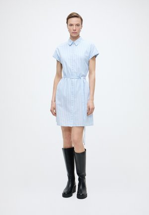 Γυναίκα που φοράει ανοιχτό μπλε ριγέ φόρεμα με κοντά μανίκια και ζώνη, συνδυασμένο με μαύρες μπότες μέχρι το γόνατο, στέκεται μπροστά σε λευκό φόντο.