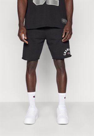 Mężczyzna ubrany w czarne szorty Champion, czarną koszulkę, białe skarpetki Champion za kostkę i białe trampki, stojący na jednolitym tle.