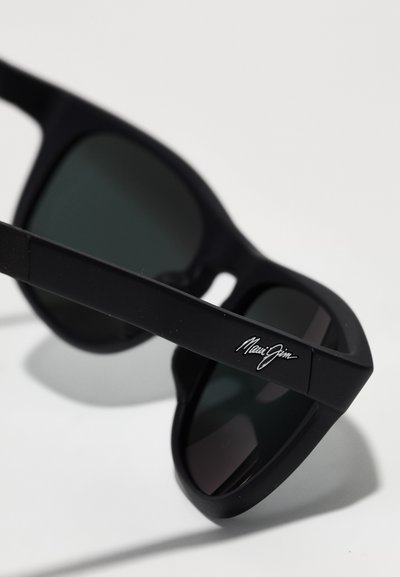 Czarne okulary przeciwsłoneczne z ciemnymi soczewkami, aerodynamiczny design i matowe wykończenie. Logo "Maui Jim" na zauszniku podkreśla oprawę.