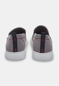 Pantofi slip-on din pânză gri, cu talpă din cauciuc alb. Prezintă o cusătură roșie și o bandă neagră de prindere etichetată "RED". Design simplu și elegant.