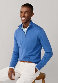 Homme souriant portant un pull bleu à fermeture éclair au niveau du cou sur une chemise rayée, pantalon blanc et ceinture marron, assis sur un tabouret en bois devant un fond uni.