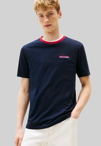 T-shirt in cotone blu navy con collo a costine rosso e bianco, con una toppa con logo sul petto. Maniche corte e vestibilità comoda.