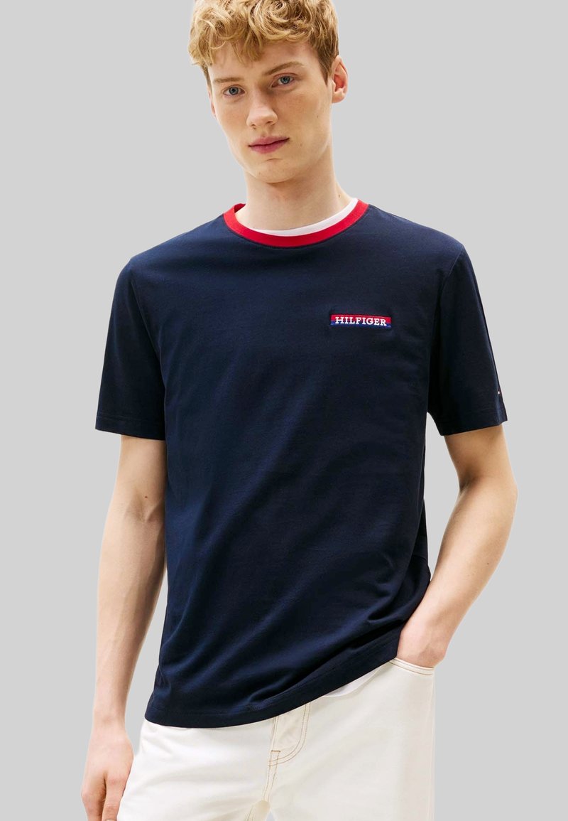 T-shirt in cotone blu navy con collo a costine rosso e bianco, con una toppa con logo sul petto. Maniche corte e vestibilità comoda.