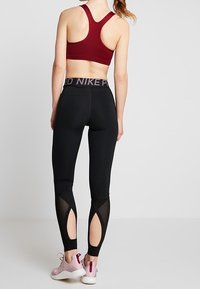 Leggings noirs avec découpes en maille au niveau du mollet, dotés d'une large ceinture avec l'inscription "NIKE PRO" en gris. Accompagnés d'un soutien-gorge de sport bordeaux.