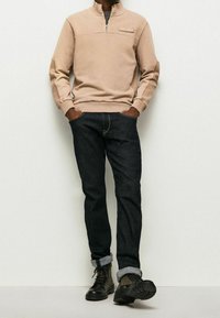 Sudadera beige con cremallera y cuello alto, combinada con pantalones de mezclilla oscuros y botas de cordones negras. Presenta puños acanalados y dobladillos en los tobillos.