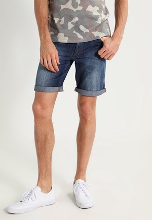 Mann trägt hochgekrempelte dunkle Jeansshorts, Camouflage-T-Shirt und weiße Sneakers, steht vor einem schlichten hellen Hintergrund.