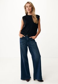 Top negro sin mangas hecho de tela de punto, combinado con unos vaqueros de pierna ancha de cintura alta en azul. Adornado con un cinturón negro. Zapatos sin tacón.