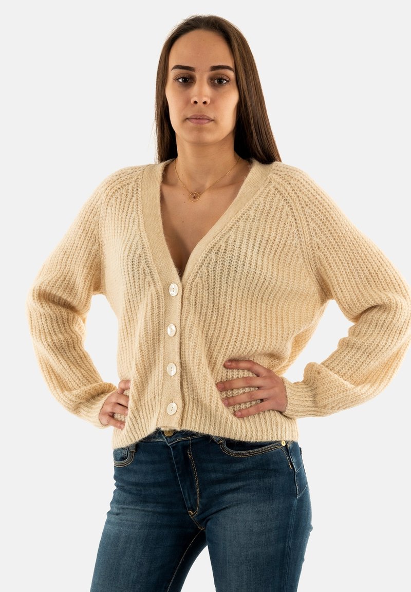 Cárdigan de punto beige con un escote en V profundo, cinco grandes botones blancos y textura acanalada. Combinado con jeans de mezclilla azul.