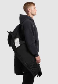 Melnā parka jaka ar oderi, kapuci, pogām un redzamu etiķeti iekšpusē. Modelis redzams no sāna, valkājot tumšu kapuci.